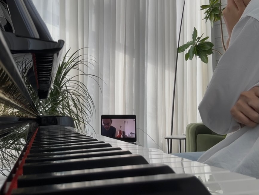Naina at the piano during an online lesson, her teacher visible on a propped-up tablet in a sunlit room