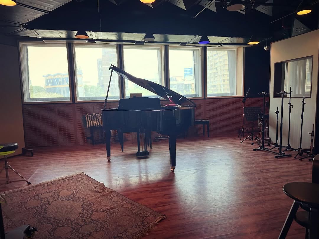 A grand piano in a window-lit Mumbai studio, with microphone stands and a rug